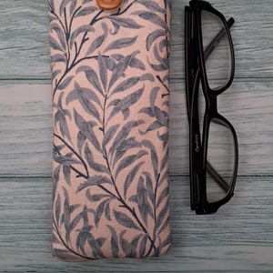 May include: A fabric eyeglasses case with a botanical leaf pattern in blue and gray on a pink background. The case has a wooden button closure and is next to a pair of black eyeglasses.