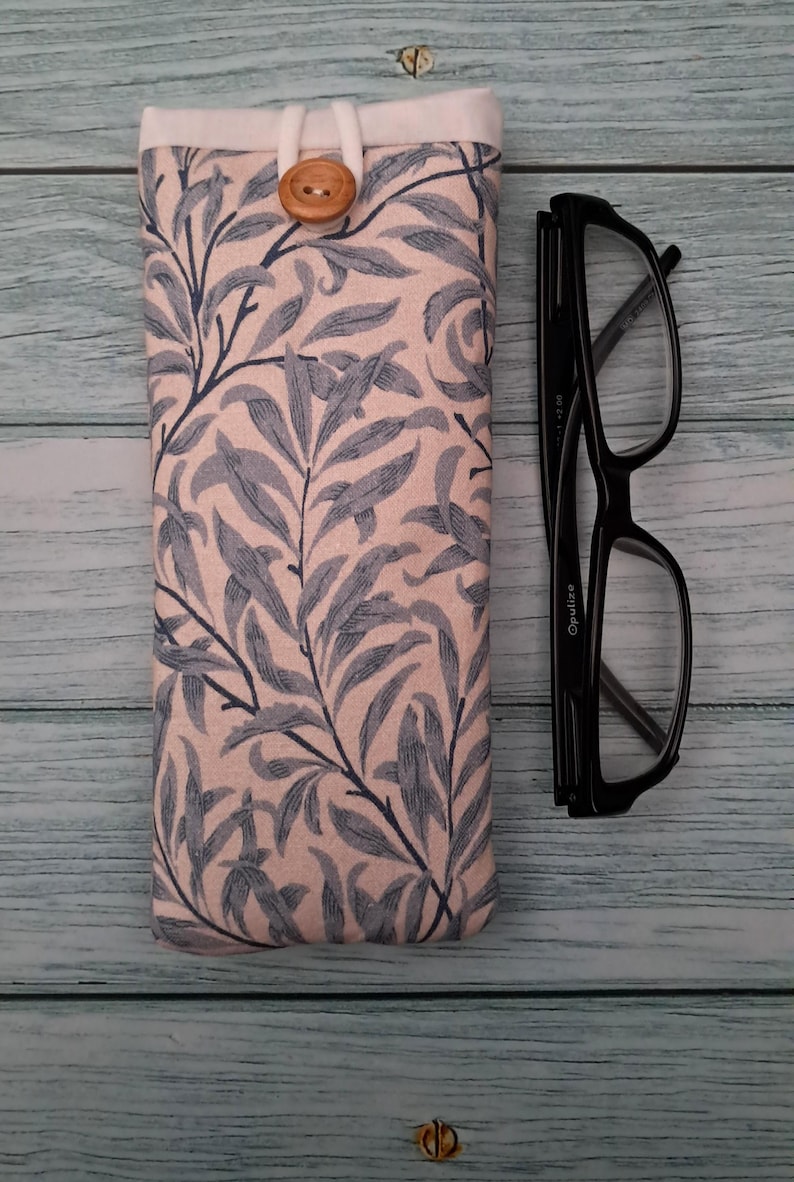 May include: A fabric eyeglasses case with a botanical leaf pattern in blue and gray on a pink background. The case has a wooden button closure and is next to a pair of black eyeglasses.