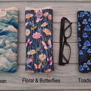 Puede incluir: Tres fundas de tela para gafas con cierre de bot&oacute;n. La primera presenta olas oce&aacute;nicas azules, la segunda muestra flores rosas y blancas con mariposas, y la tercera muestra setas azules sobre un fondo negro. Se incluyen unas gafas negras.