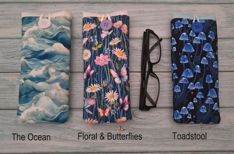 Puede incluir: Tres fundas de tela para gafas con cierre de bot&oacute;n. La primera presenta olas oce&aacute;nicas azules, la segunda muestra flores rosas y blancas con mariposas, y la tercera muestra setas azules sobre un fondo negro. Se incluyen unas gafas negras.