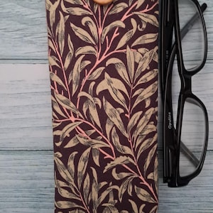 May include: A fabric glasses case with a botanical pattern in shades of brown, beige, and pink. The case has a button closure and a white top. Black eyeglasses are next to the case.