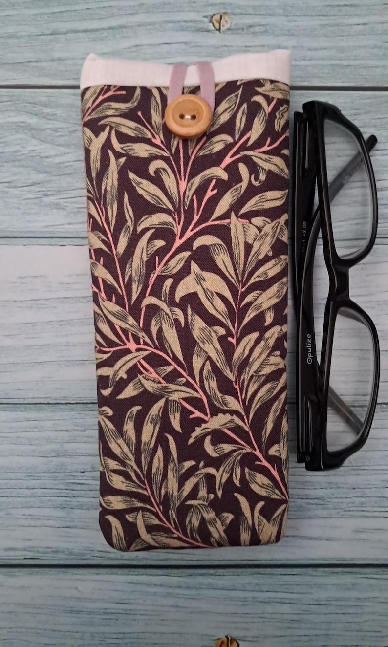 May include: A fabric glasses case with a botanical pattern in shades of brown, beige, and pink. The case has a button closure and a white top. Black eyeglasses are next to the case.