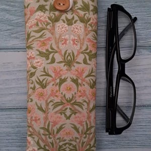 May include: A floral eyeglasses case with a button closure next to a pair of black eyeglasses. The case features a pink and green floral pattern on a light beige background. The glasses have a rectangular frame.