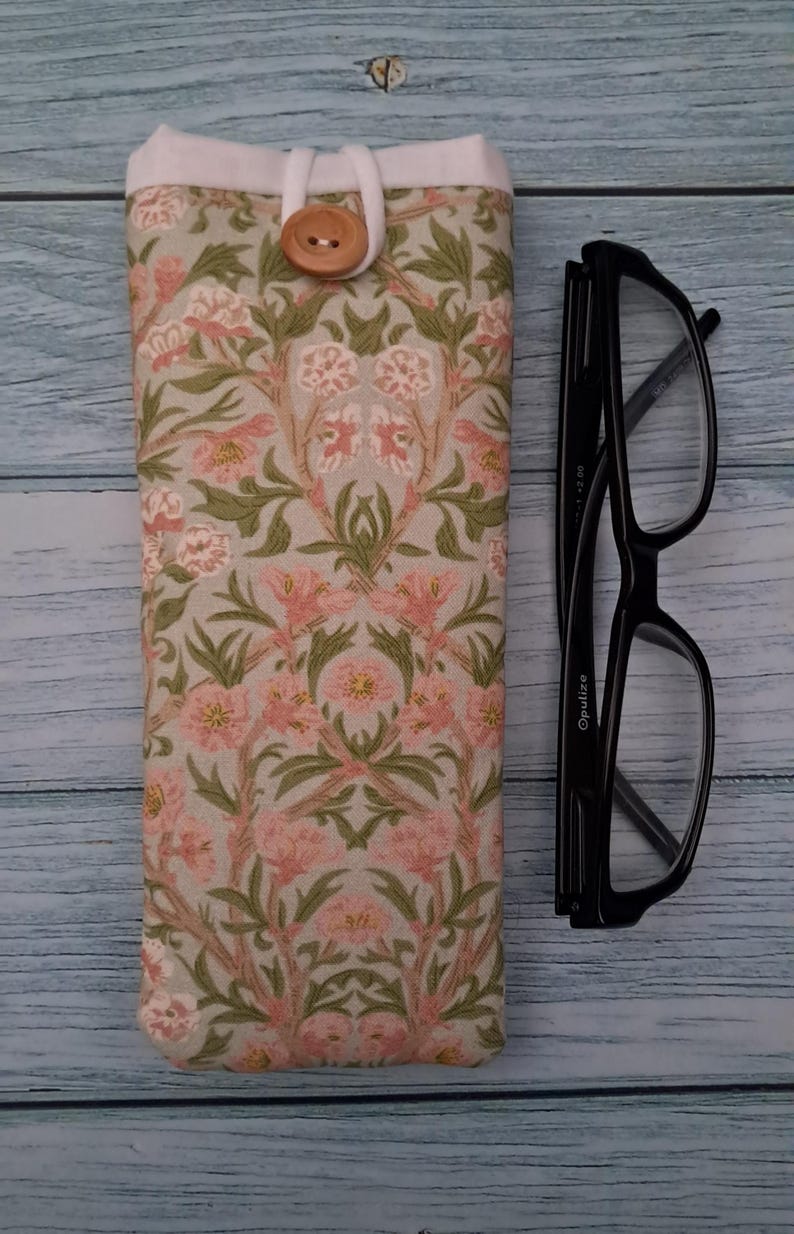 May include: A floral eyeglasses case with a button closure next to a pair of black eyeglasses. The case features a pink and green floral pattern on a light beige background. The glasses have a rectangular frame.