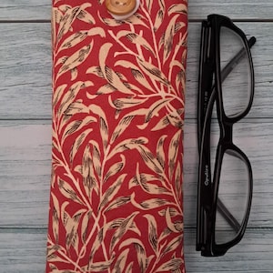 May include: A red and cream floral print glasses case with a wooden button closure. Next to the case is a pair of black eyeglasses. The case is rectangular and has a white top.