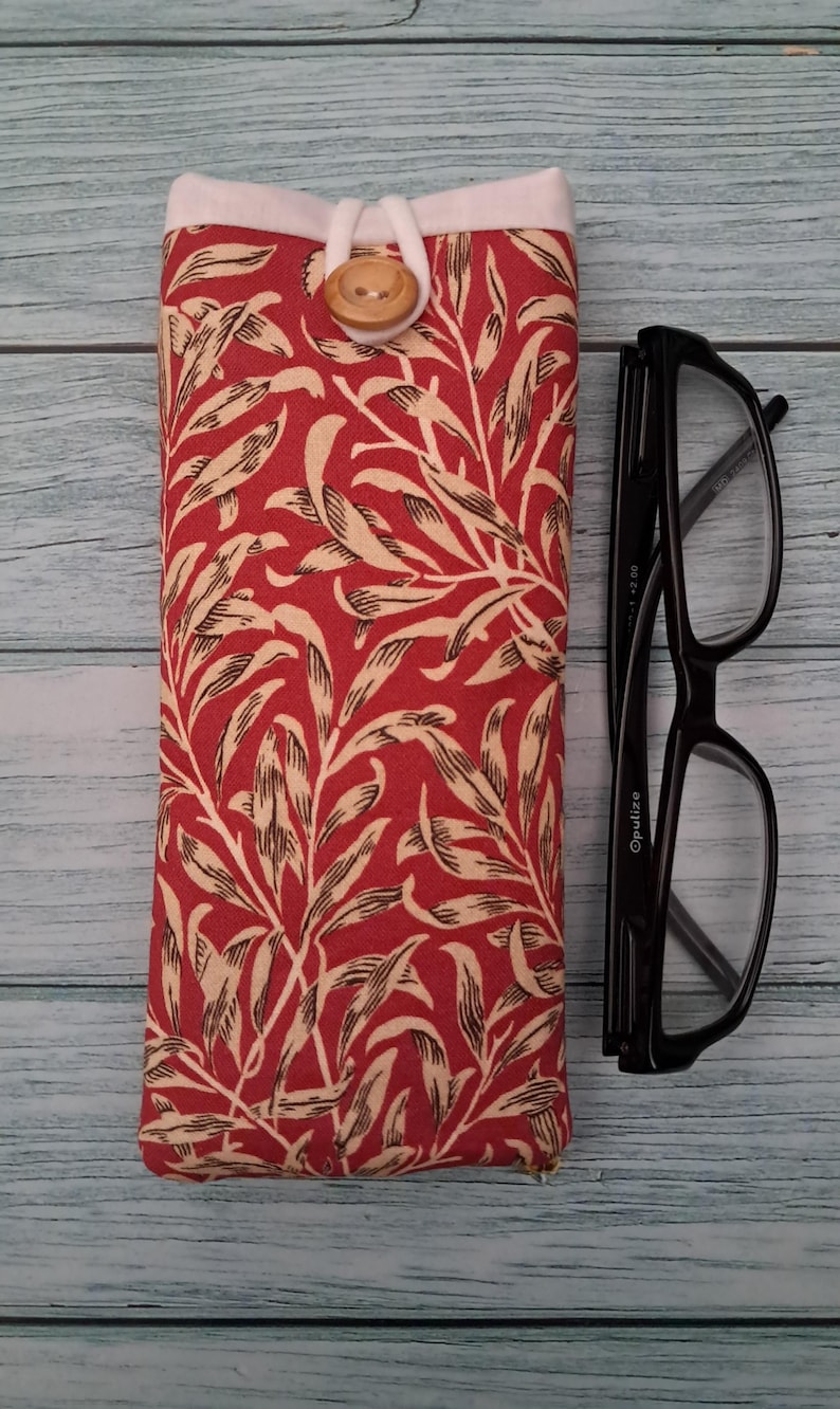 May include: A red and cream floral print glasses case with a wooden button closure. Next to the case is a pair of black eyeglasses. The case is rectangular and has a white top.