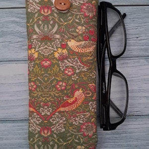 May include: A green eyeglasses case with a floral and bird pattern, a white top, and a wooden button closure. Black eyeglasses are next to the case. The case is approximately 7 inches long.