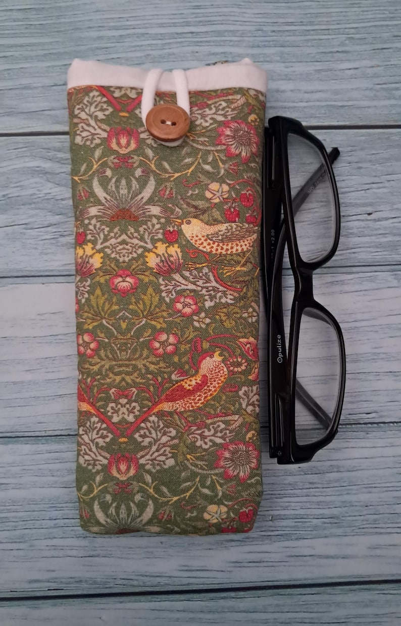 May include: A green eyeglasses case with a floral and bird pattern, a white top, and a wooden button closure. Black eyeglasses are next to the case. The case is approximately 7 inches long.