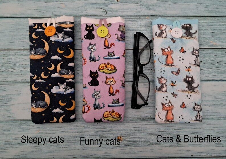 Puede incluir: Tres fundas de tela para gafas con cierre de bot&oacute;n. La primera es azul oscuro con lunas amarillas y gatos durmiendo. La segunda es morado claro con varios gatos. La tercera es azul claro con gatos y mariposas.