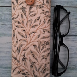 May include: A fabric eyeglasses case with a botanical leaf pattern in beige, brown, and black. The case is closed with a wooden button and loop. Black eyeglasses are next to the case.
