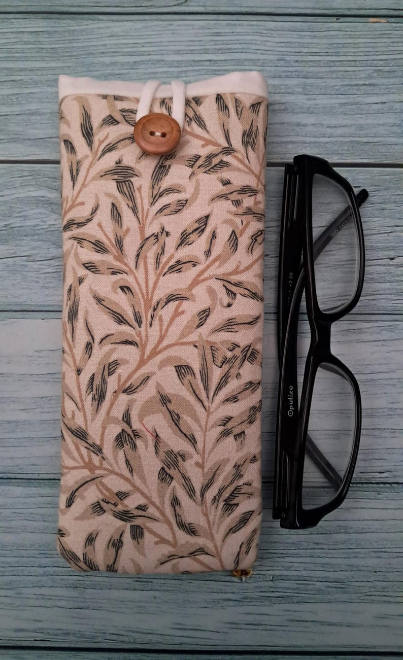 May include: A fabric eyeglasses case with a botanical leaf pattern in beige, brown, and black. The case is closed with a wooden button and loop. Black eyeglasses are next to the case.
