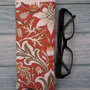 May include: A floral patterned glasses case in shades of red, white, and green, with a wooden button closure. Next to it are black-framed eyeglasses. The case features a botanical design.