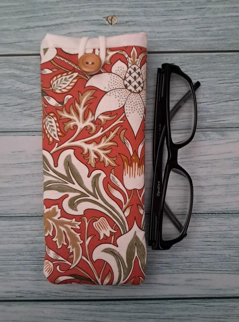 May include: A floral patterned glasses case in shades of red, white, and green, with a wooden button closure. Next to it are black-framed eyeglasses. The case features a botanical design.