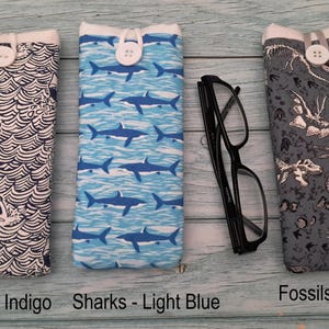 Puede incluir: Tres estuches rectangulares de tela para gafas con cierre de bot&oacute;n. El primero presenta un patr&oacute;n de tiburones y olas azul oscuro, el segundo un patr&oacute;n de tiburones azul claro y el tercero un estampado de f&oacute;siles grises. Unas gafas negras.