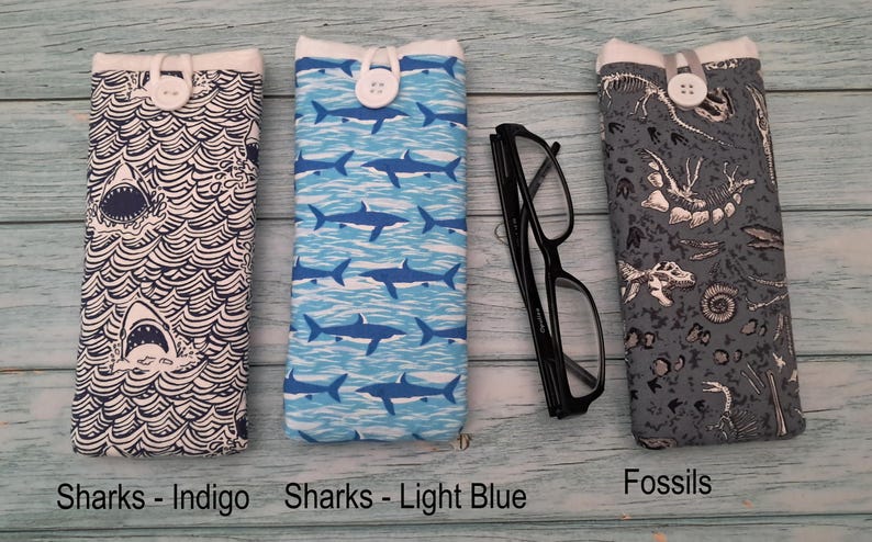 Puede incluir: Tres estuches rectangulares de tela para gafas con cierre de bot&oacute;n. El primero presenta un patr&oacute;n de tiburones y olas azul oscuro, el segundo un patr&oacute;n de tiburones azul claro y el tercero un estampado de f&oacute;siles grises. Unas gafas negras.