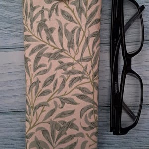 May include: A fabric eyeglasses case with a botanical print in green and gray on a pink background. The case has a white top and a wooden button closure. Black eyeglasses are next to the case.