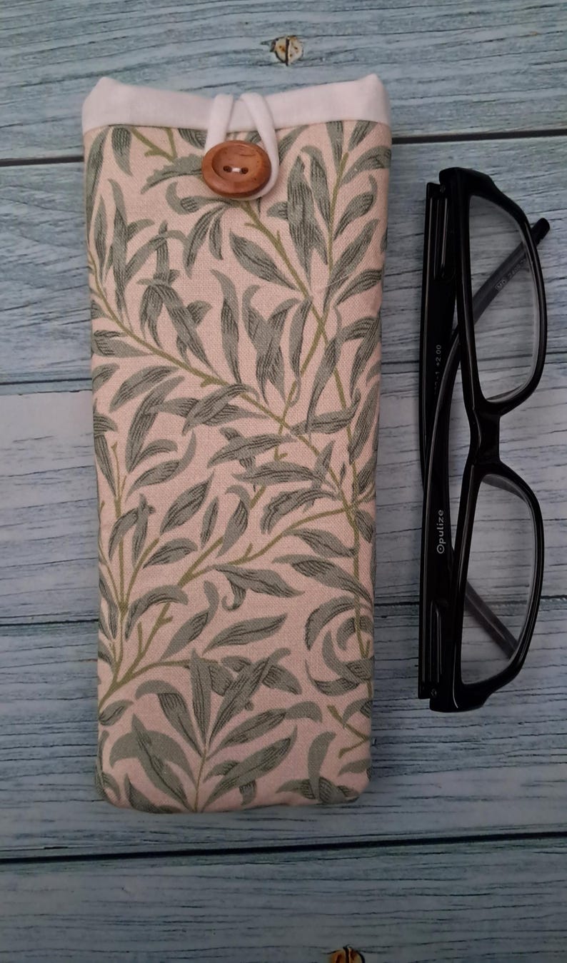 May include: A fabric eyeglasses case with a botanical print in green and gray on a pink background. The case has a white top and a wooden button closure. Black eyeglasses are next to the case.