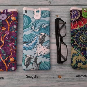 Puede incluir: Tres estuches rectangulares de tela con cierre de bot&oacute;n. El estuche izquierdo presenta un patr&oacute;n de pavo real y flores sobre fondo morado con un bot&oacute;n morado. El central muestra gaviotas sobre olas azules con un bot&oacute;n blanco. El derecho tiene un estampado de f&oacute;siles de amonites coloridos con un bot&oacute;n verde. Unas gafas negras se sit&uacute;an entre el estuche central y el derecho.