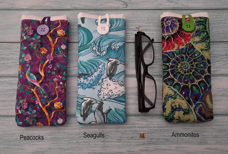 Puede incluir: Tres estuches rectangulares de tela con cierre de bot&oacute;n. El estuche izquierdo presenta un patr&oacute;n de pavo real y flores sobre fondo morado con un bot&oacute;n morado. El central muestra gaviotas sobre olas azules con un bot&oacute;n blanco. El derecho tiene un estampado de f&oacute;siles de amonites coloridos con un bot&oacute;n verde. Unas gafas negras se sit&uacute;an entre el estuche central y el derecho.