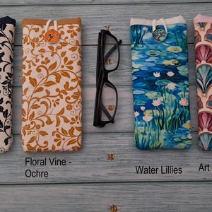 Puede incluir: Cuatro fundas de tela para gafas con cierre de bot&oacute;n, etiquetadas como Floral Vine-Navy, Floral Vine-Ochre, Water Lillies y Art Deco Shells, dispuestas junto a unas gafas de montura negra. Las fundas presentan patrones distintos en tonos azul marino, ocre, azul y rosa.