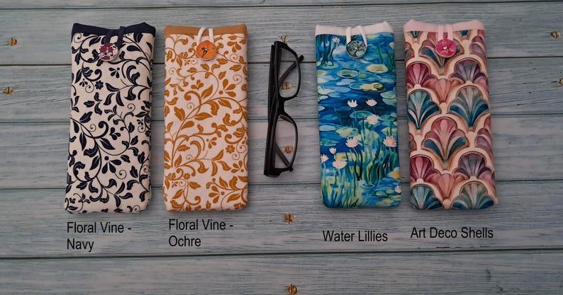 Puede incluir: Cuatro fundas de tela para gafas con cierre de bot&oacute;n, etiquetadas como Floral Vine-Navy, Floral Vine-Ochre, Water Lillies y Art Deco Shells, dispuestas junto a unas gafas de montura negra. Las fundas presentan patrones distintos en tonos azul marino, ocre, azul y rosa.