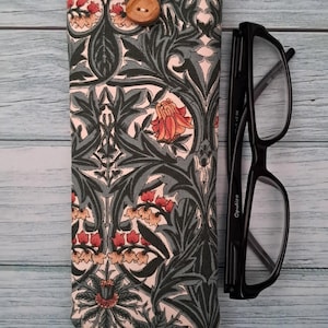 May include: A patterned glasses case with a floral design in shades of green, red, and cream, secured with a wooden button. Black eyeglasses are placed next to the case. The case is approximately 7 inches long.
