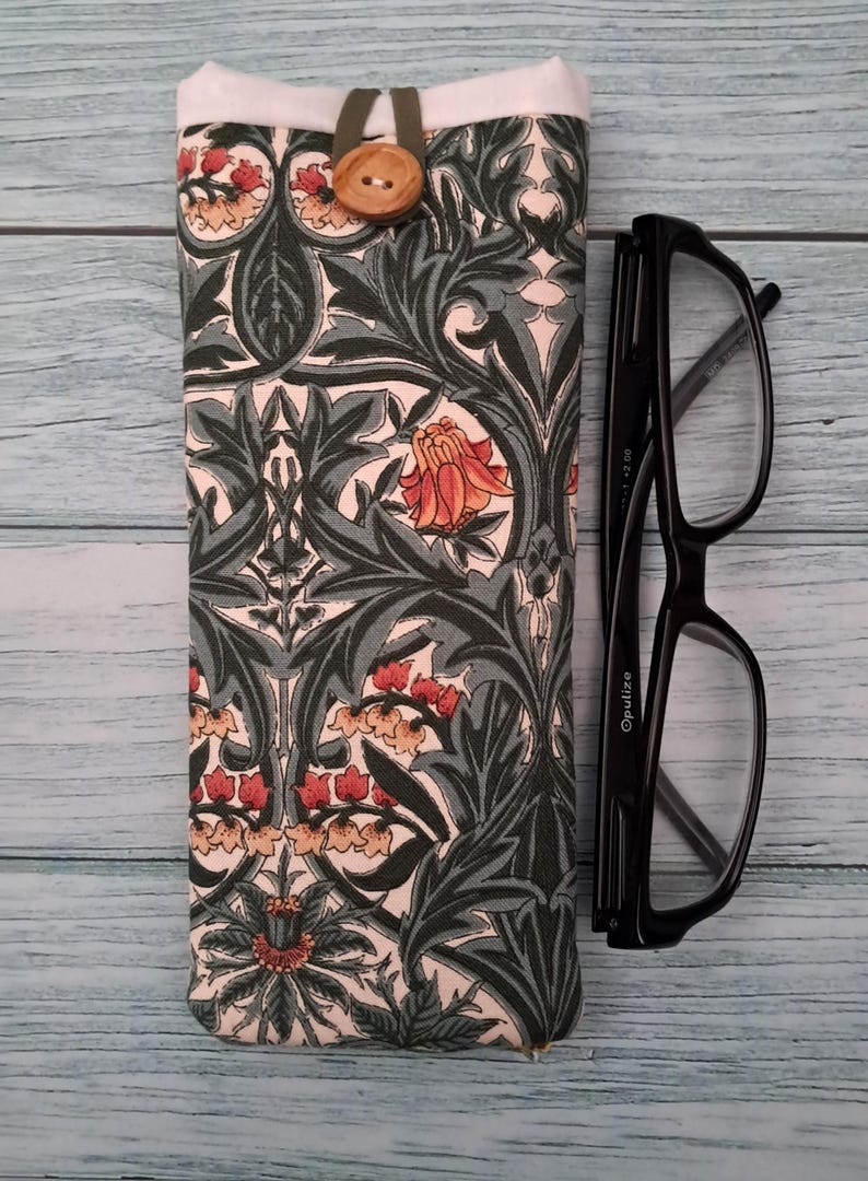 May include: A patterned glasses case with a floral design in shades of green, red, and cream, secured with a wooden button. Black eyeglasses are placed next to the case. The case is approximately 7 inches long.