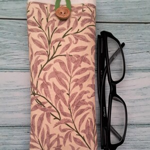 May include: A fabric eyeglasses case with a botanical print in shades of beige, mauve, and green. The case is closed with a wooden button and a green fabric loop. Black eyeglasses are next to the case.