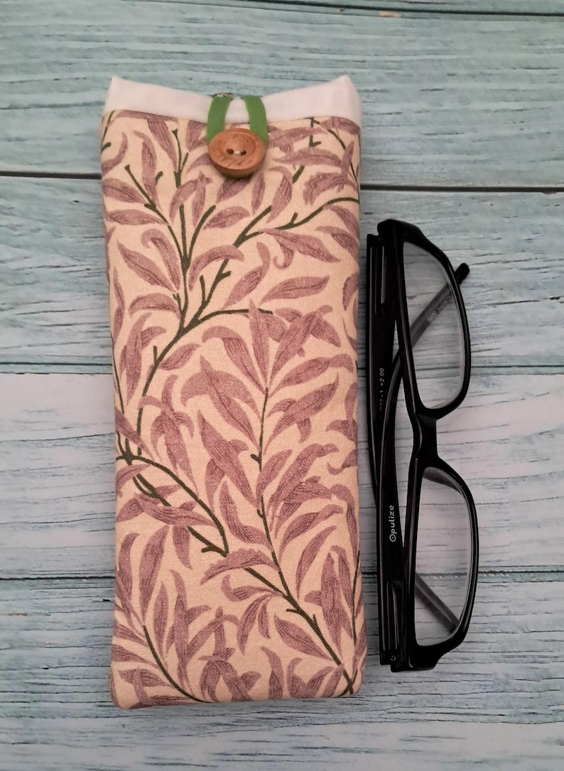 May include: A fabric eyeglasses case with a botanical print in shades of beige, mauve, and green. The case is closed with a wooden button and a green fabric loop. Black eyeglasses are next to the case.