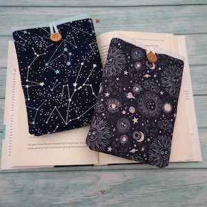 May include: Two fabric book sleeves with celestial patterns. One shows constellations on a navy background, the other has suns, moons, and planets. Each has a light blue trim and a wooden button closure.