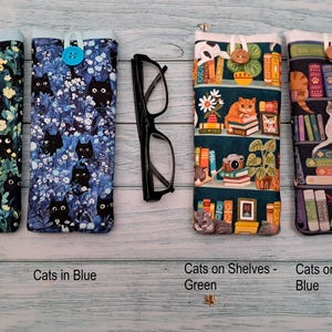 Puede incluir: Cuatro estuches rectangulares de tela con cierres de bot&oacute;n y un par de gafas negras. Dos estuches presentan patrones de gatos negros sobre fondos verde y azul. Dos estuches presentan patrones de gatos y estanter&iacute;as de libros.