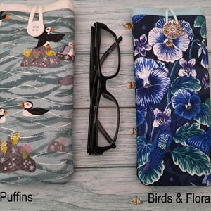 Puede incluir: Dos fundas de tela para gafas y unas gafas de pl&aacute;stico negro. La funda izquierda tiene un fondo azul claro con frailecillos y rocas. La funda derecha tiene un fondo azul oscuro con flores azules y p&aacute;jaros. Ambas fundas tienen cierres de bot&oacute;n.