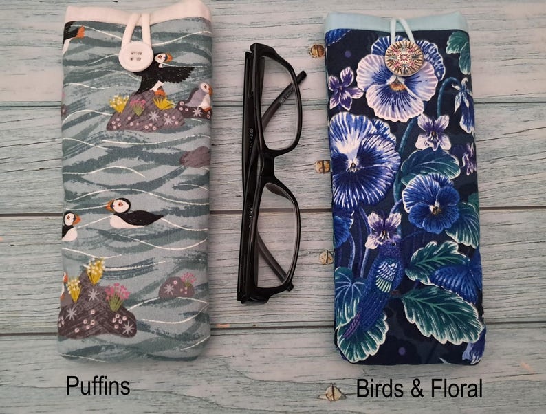 Puede incluir: Dos fundas de tela para gafas y unas gafas de pl&aacute;stico negro. La funda izquierda tiene un fondo azul claro con frailecillos y rocas. La funda derecha tiene un fondo azul oscuro con flores azules y p&aacute;jaros. Ambas fundas tienen cierres de bot&oacute;n.