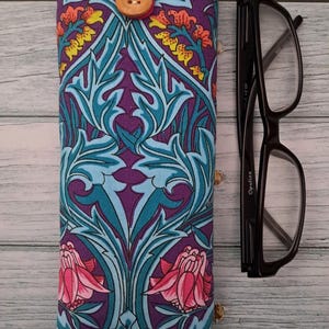May include: A fabric glasses case with a floral pattern in purple, teal, and pink. The case has a wooden button closure and a pink strap. Black eyeglasses are next to the case.