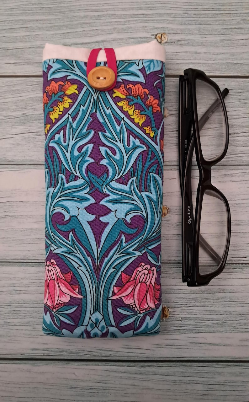 May include: A fabric glasses case with a floral pattern in purple, teal, and pink. The case has a wooden button closure and a pink strap. Black eyeglasses are next to the case.