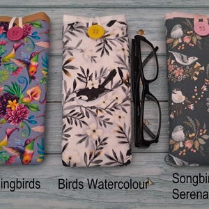 Puede incluir: Tres fundas de tela para gafas con cierre de bot&oacute;n. La funda de la izquierda presenta colibr&iacute;es y flores, la central tiene un patr&oacute;n de aves en blanco y negro con un bot&oacute;n amarillo, y la derecha muestra p&aacute;jaros cantores y motivos florales.