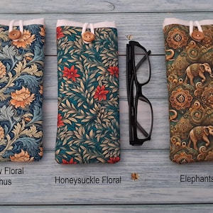 Puede incluir: Tres fundas de tela para gafas con cierre de bot&oacute;n. La primera presenta un patr&oacute;n floral de acanto amarillo, la segunda un estampado floral de madreselva y la tercera un motivo de elefantes. Unas gafas negras se sit&uacute;an entre las fundas.