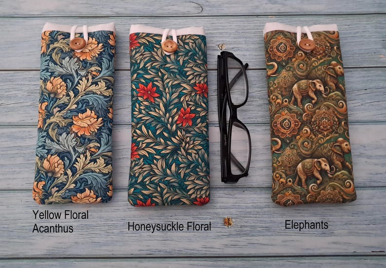 Puede incluir: Tres fundas de tela para gafas con cierre de bot&oacute;n. La primera presenta un patr&oacute;n floral de acanto amarillo, la segunda un estampado floral de madreselva y la tercera un motivo de elefantes. Unas gafas negras se sit&uacute;an entre las fundas.