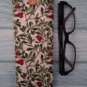 May include: A fabric eyeglasses case with a botanical pattern of green leaves and red berries on a light gray background. The case has a white top with a wooden button closure. Black eyeglasses are next to the case.