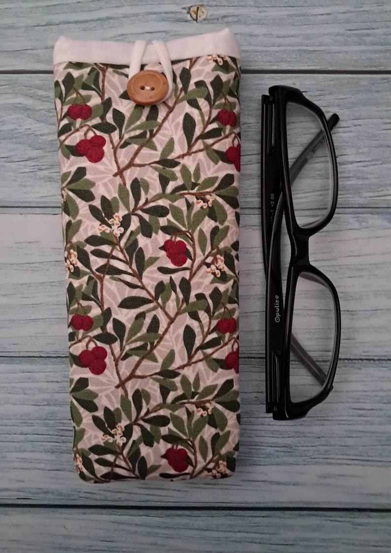 May include: A fabric eyeglasses case with a botanical pattern of green leaves and red berries on a light gray background. The case has a white top with a wooden button closure. Black eyeglasses are next to the case.