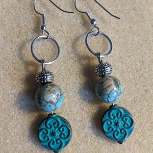 May include: A pair of dangle earrings with turquoise and silver beads. The earrings feature a silver hoop, a silver bead, a turquoise bead, and a turquoise charm with a floral design.