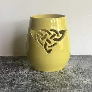 May include: A pale yellow ceramic candle holder with a rounded, tapered shape. The front features a cutout design of a traditional triquetra Celtic knot, allowing light to pass through the open spaces of the intricate, interlaced pattern.