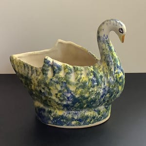 May include: A ceramic swan-shaped planter featuring a mottled glaze in shades of blue, yellow, and white. The swan has a long, curved neck, a small orange beak, and a black eye. The interior of the vessel is a smooth, off-white cream color.
