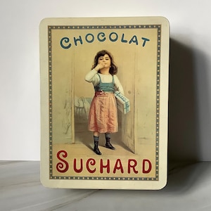 May include: Rectangular vintage tin featuring a young girl eating chocolate. The tin has the words "CHOCOLAT SUCHARD" in green and red lettering. The girl is wearing a white top, pink skirt, and black shoes. The background is a light brown color.