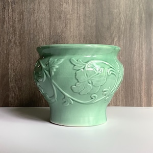 May include: A light mint green ceramic planter featuring a raised relief pattern of flowers and swirling vines around the exterior. The vessel has a wide, rounded body that tapers toward a circular base and a smooth, glossy finish throughout.
