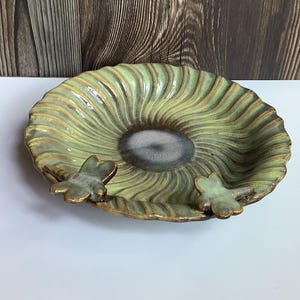 May include: A round ceramic dish featuring a light green, spiraling, ribbed pattern radiating from a dark gray center. Two small, sculpted dragonfly figures are attached to the scalloped rim, finished in a metallic bronze glaze.