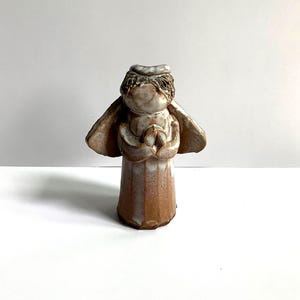 May include: A ceramic angel figurine with a light brown and cream glaze. The angel has outstretched wings and is holding its hands together in a prayerful pose. The figurine is standing on a white surface.