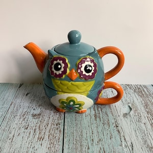 May include: A whimsical owl-shaped teapot with a teal body, orange handle and spout, and a teal lid. The owl's face features large purple and white eyes, an orange beak, and a yellow and green chest. The teapot is decorated with floral patterns.