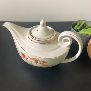 May include: Cream-colored ceramic teapot with a gold rim and a decorative floral pattern in orange and yellow. The teapot has a curved handle and a matching lid. A metal container with green packets and a wooden lid are next to the teapot.