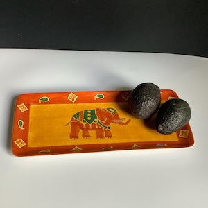 May include: A rectangular serving tray with an orange border and a yellow center. The tray features a decorative elephant design in the center and two avocados. The tray is approximately 12 inches long and 6 inches wide.